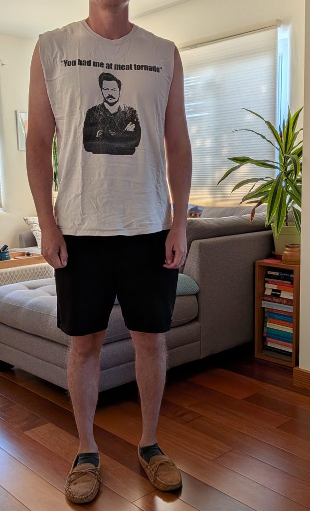 A man, whose head is not visible, standing in a living room and wearing a sleeveless t-shirt with an image of Ron Swanson on it, and the text, 