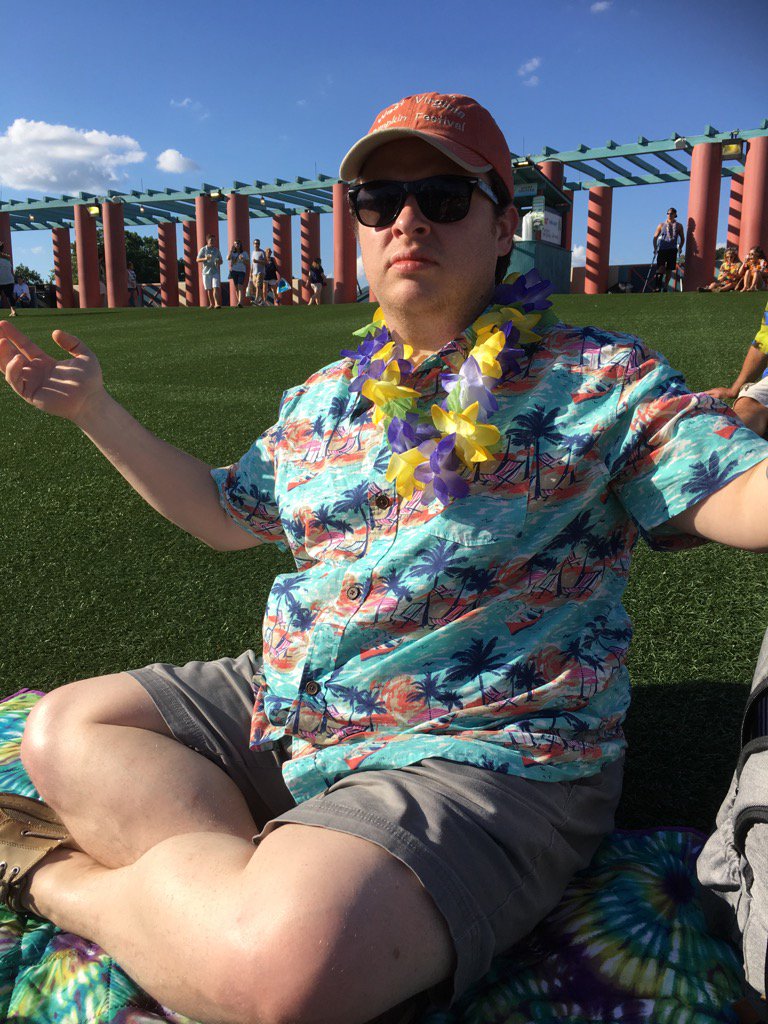 A man wearing an aloha shirt and ballcap, sitting criss cross applesauce on grass