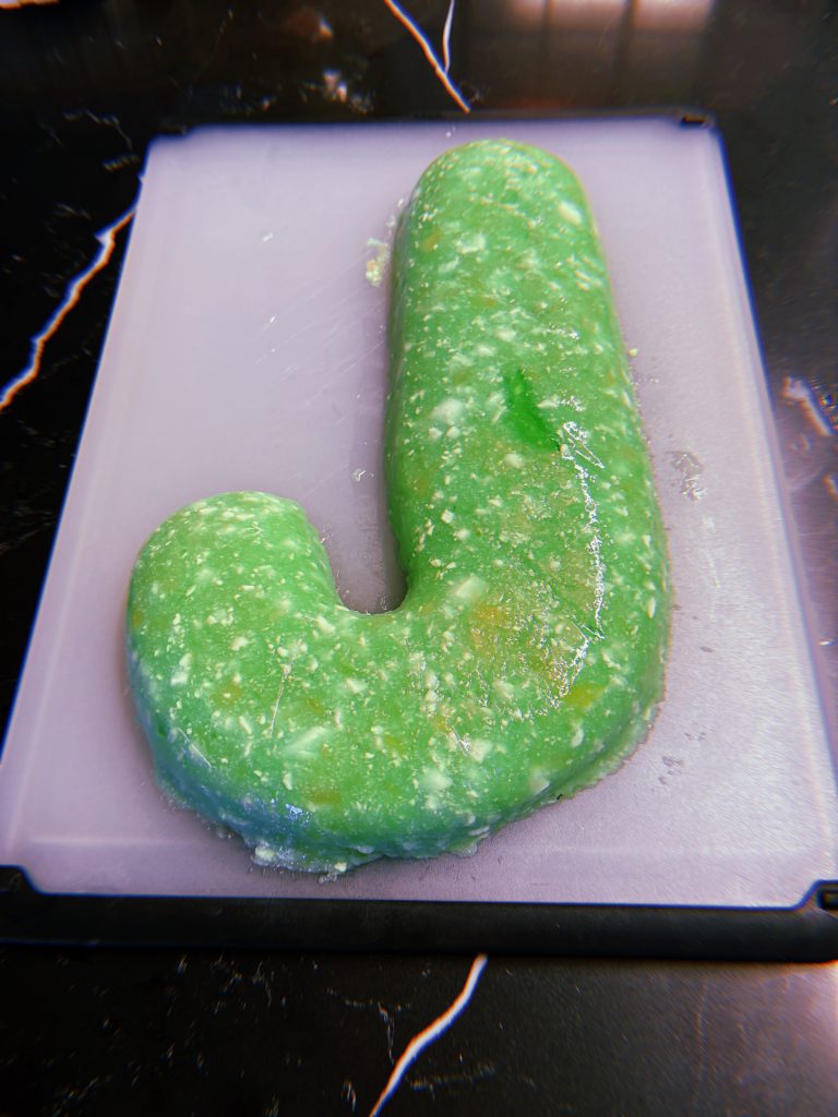 A light green jello mold in the shape of a J. The Jello has bits of cottage cheese and crushed pineapple distributed throughout.