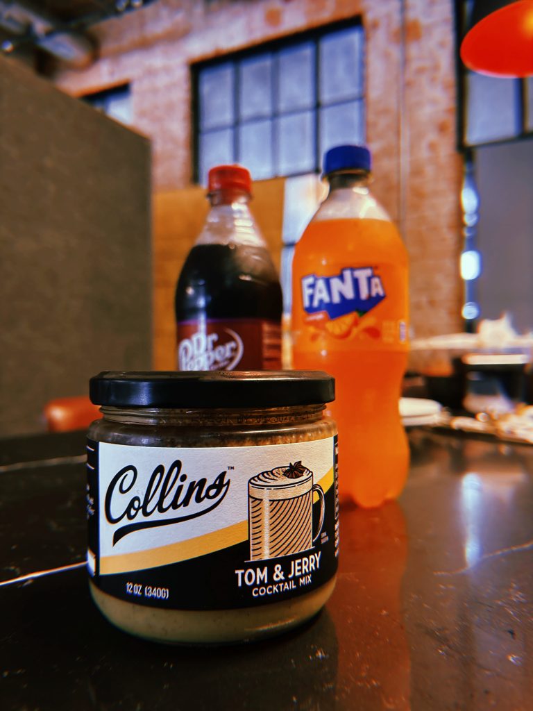A jar of Collins brand Tom and Jerry mix in the foreground. Blackberry Dr Pepper and Orange Fanta in the background
