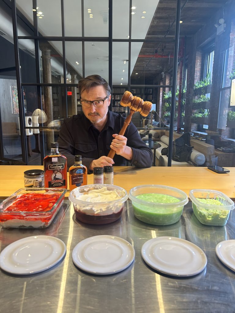 A man holding a large gavel, standing behind a counter with four jello salads on it.