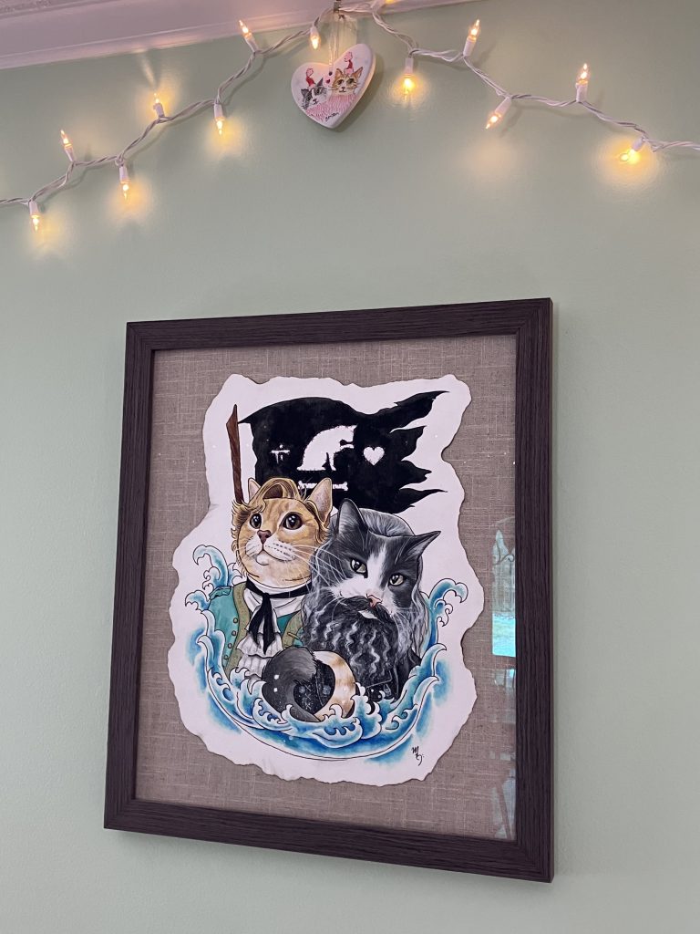 a framed piece of art depicting two cats with a cat pirate glad behind them