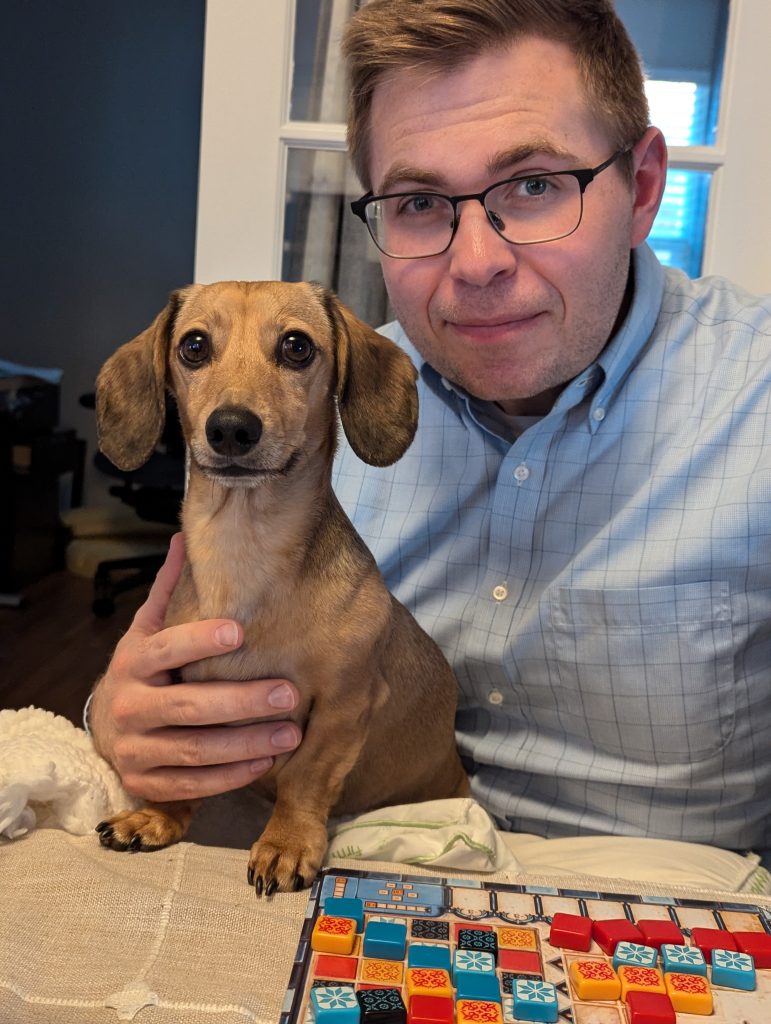 A dachshund sitting on a man's lap in front of a table with the board game AZUL on it