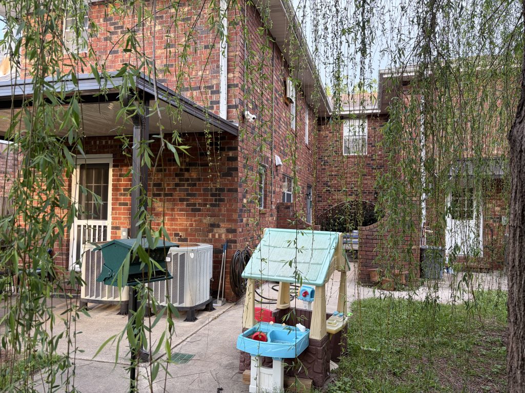 A brick house's backyard, as seen through willow tree branches. In the foreground is a children's play house