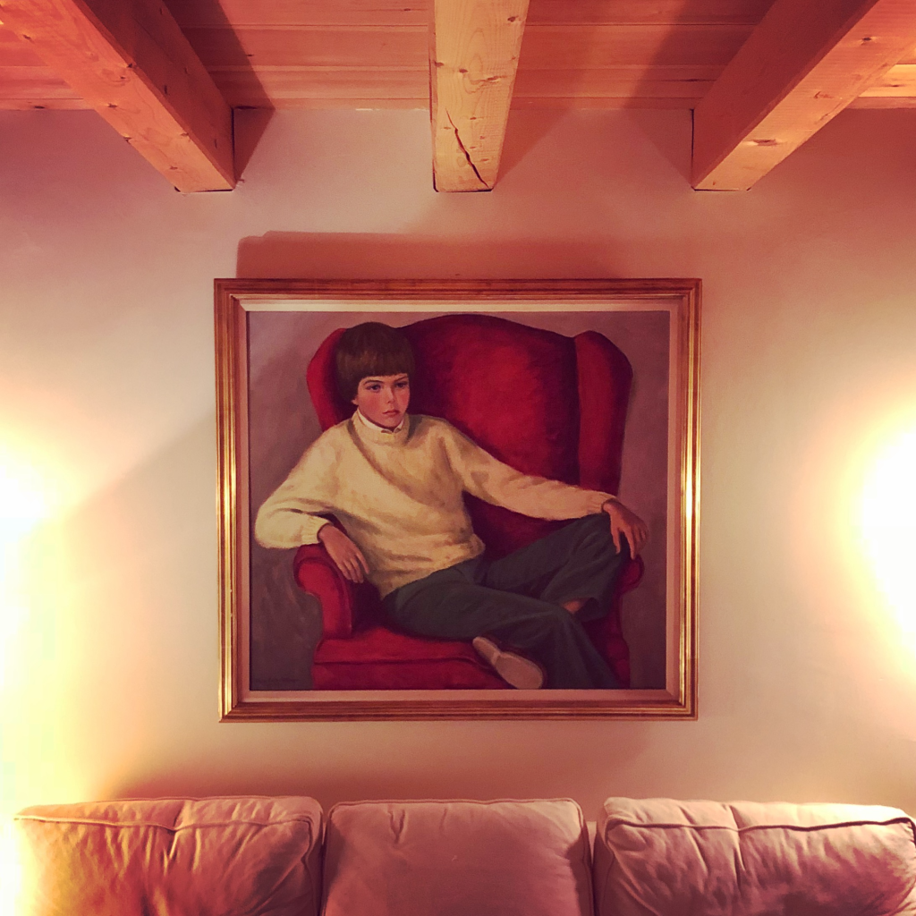 A painting hung on a wall above a sofa. The subject of the painting is a sad looking boy sitting on a red armchair