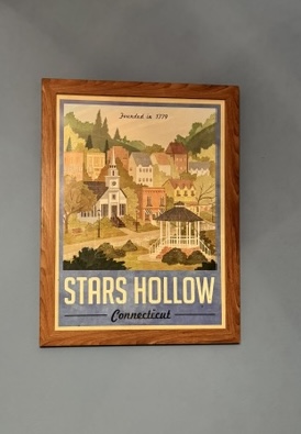 A framed poster on a wall in the style of a tourism poster for Stars Hollow. An illustration of the fictional New England Town takes up most of the space with 