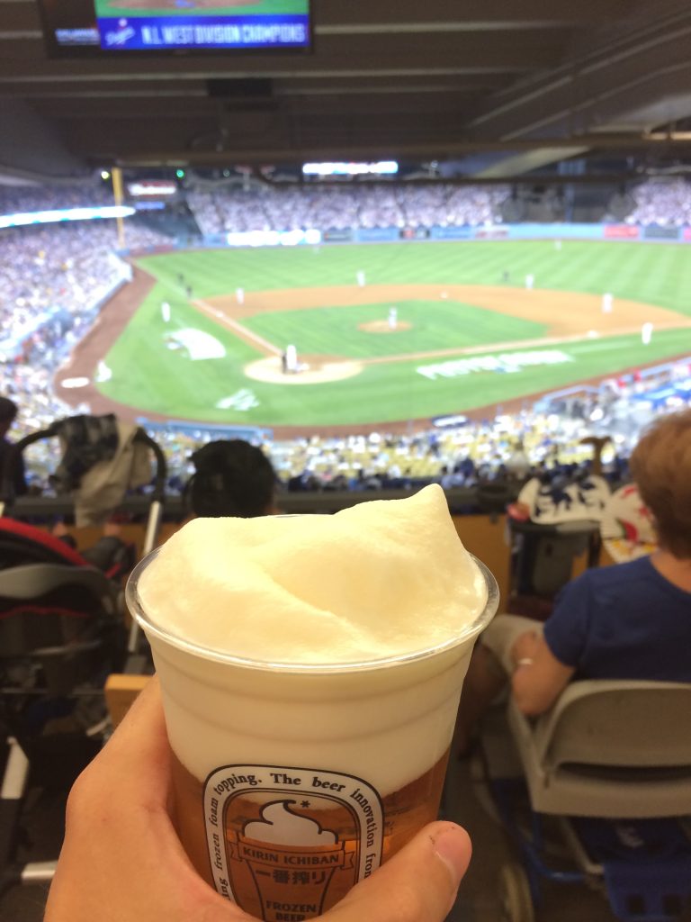 In the background is the baseball diamond at Dodger Stadium. In the foreground there is a hand holding a very foamy beer