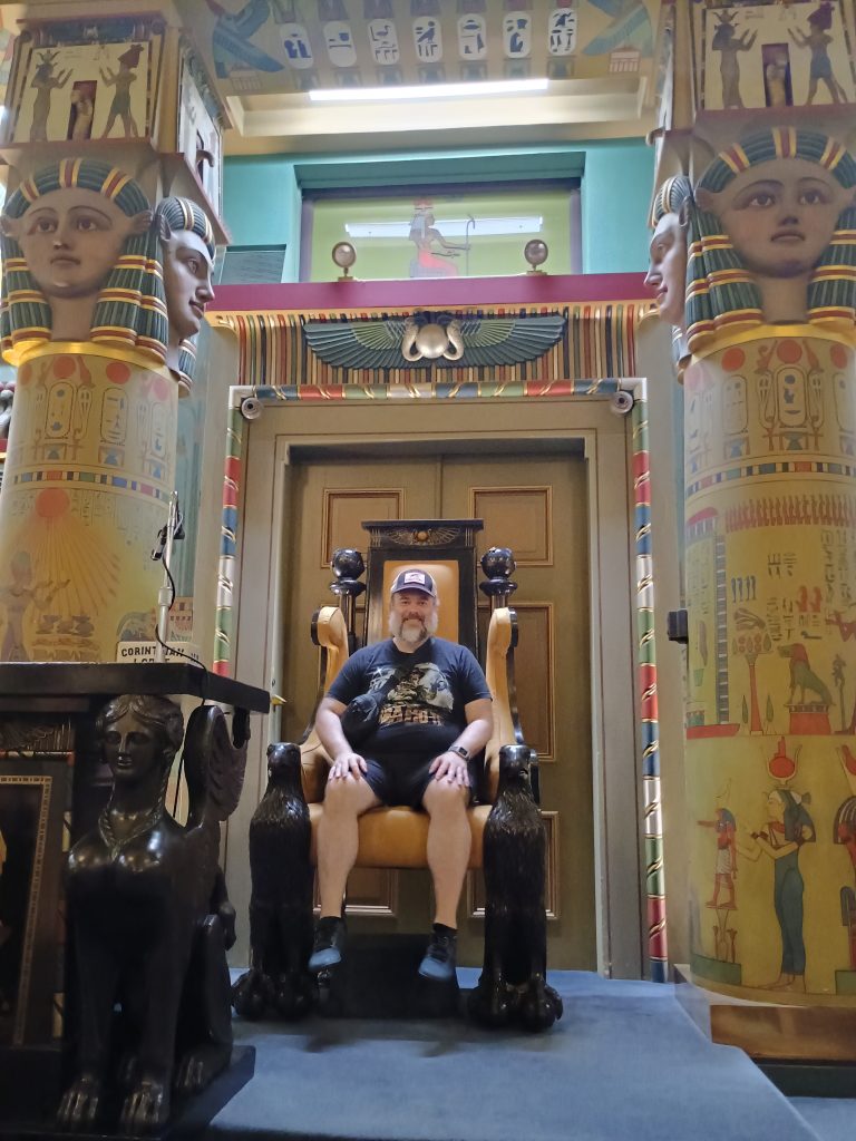 A man sitting in a large chair in an ornately decorated room wearing a crossbody bag